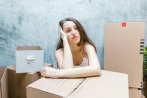 A young woman sitting among cardboard boxes looking stressed, possibly regretting hiring a moving company without proper research.