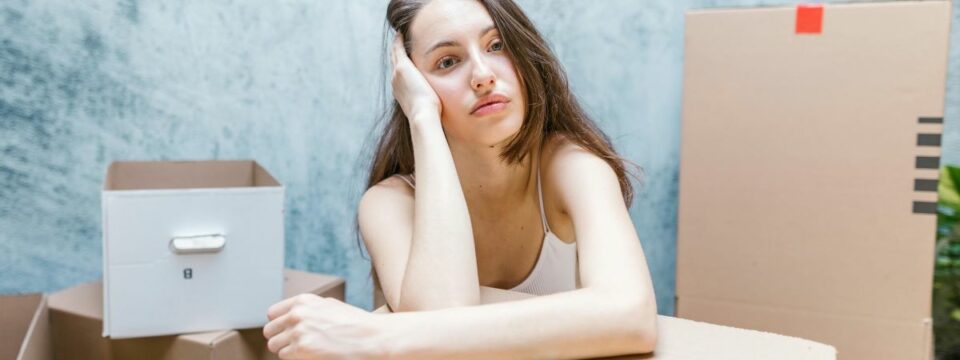 A young woman sitting among cardboard boxes looking stressed, possibly regretting hiring a moving company without proper research.