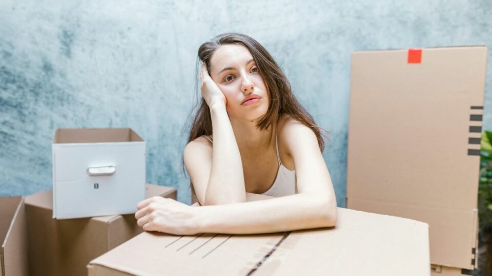 A young woman sitting among cardboard boxes looking stressed, possibly regretting hiring a moving company without proper research.