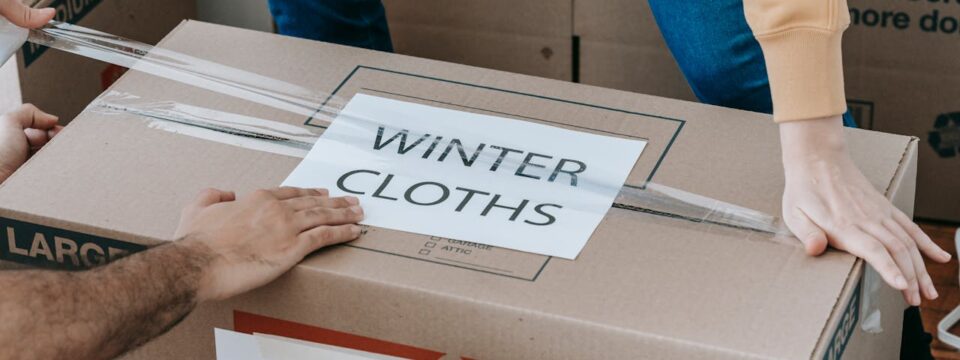 Two people sealing a moving box labeled 'Winter Clothes' as part of smart labeling systems for an organized unpacking process.