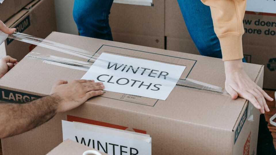 Two people sealing a moving box labeled 'Winter Clothes' as part of smart labeling systems for an organized unpacking process.