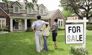 A trio of individuals poses in front of a house displaying a "Sold" sign, marking the completion of their real estate deal.