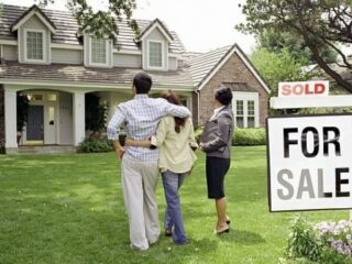 A trio of individuals poses in front of a house displaying a "Sold" sign, marking the completion of their real estate deal.